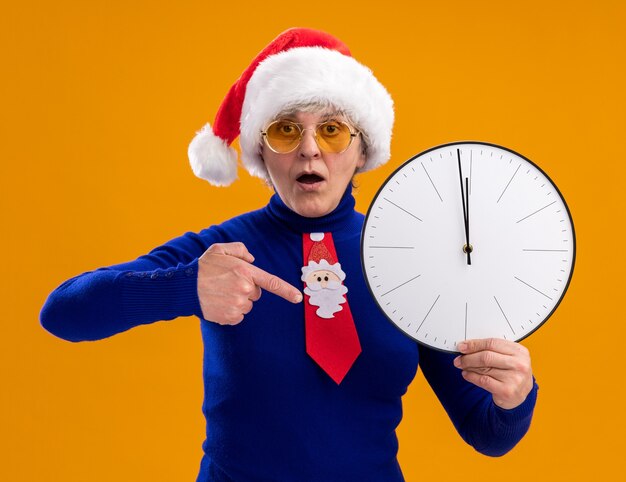 Surprised elderly woman in sun glasses with santa hat and santa tie holding and pointing at clock isolated on orange wall with copy space