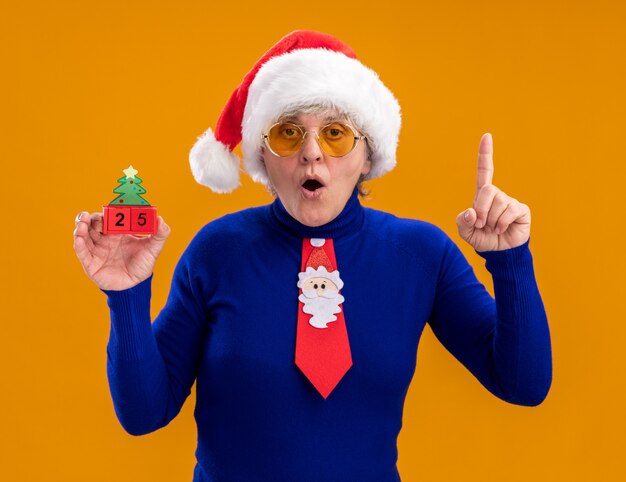 Surprised elderly woman in sun glasses with santa hat and santa tie holds christmas tree ornament and points up isolated on orange wall with copy space