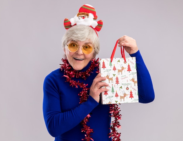 Surprised elderly woman in sun glasses with santa headband and garland around neck holding paper gift bag 