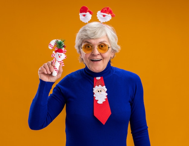 Free photo surprised elderly woman in sun glasses with santa headband and santa tie holding candy cane isolated on orange background with copy space