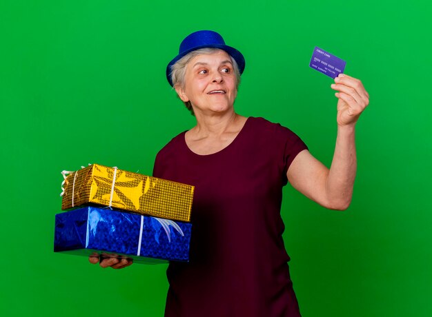 Surprised elderly woman wearing party hat holds gift boxes and looks at credit card on green