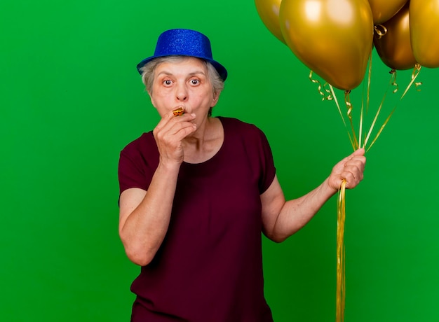 Surprised elderly woman wearing party hat holds helium balloons blowing whistle on green