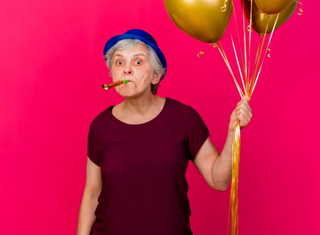 Surprised elderly woman wearing party hat holds helium balloons blowing whistle on pink