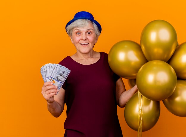 Surprised elderly woman wearing party hat holds helium balloons and money isolated on orange wall with copy space