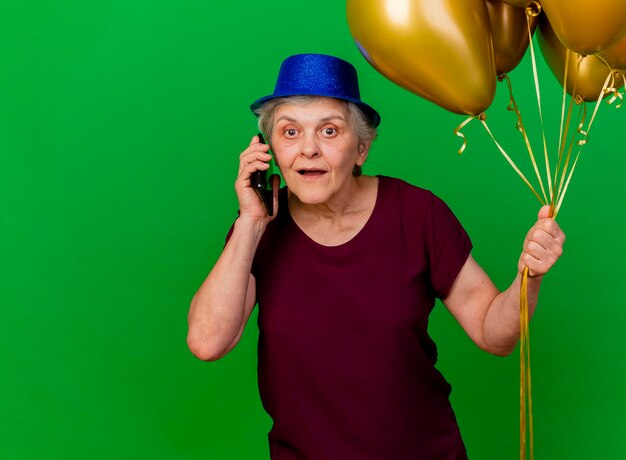 Surprised elderly woman wearing party hat holds helium balloons talking on phone on green