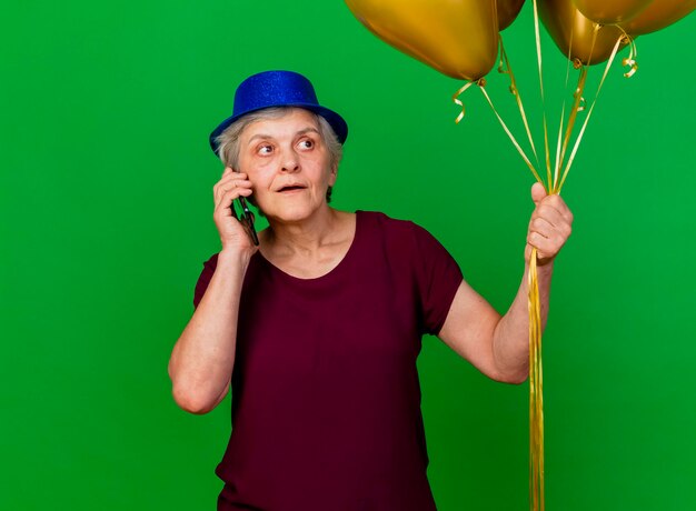 パーティーハットをかぶった驚いた年配の女性は、ヘリウム気球を持って、緑の側を見て電話で話します