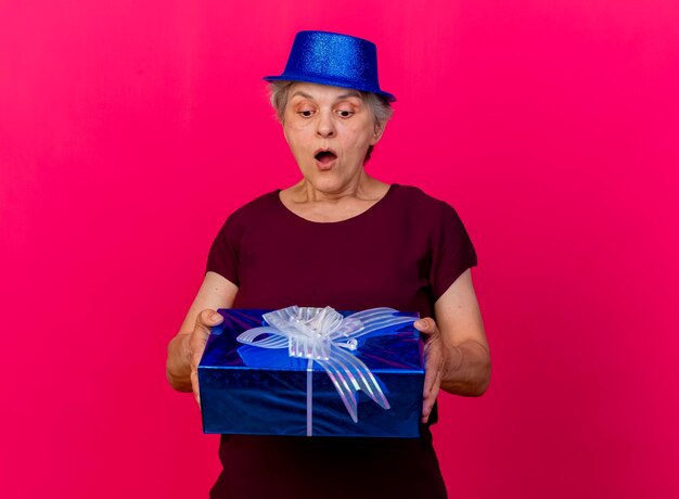 Surprised elderly woman wearing party hat holds and looks at gift box on pink