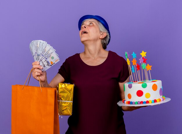 Surprised elderly woman wearing party hat holds money gift box paper shopping bag and birthday cake isolated on purple wall with copy space