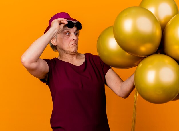 Surprised elderly woman wearing party hat holds sun glasses standing and looking at helium balloons isolated on orange wall with copy space