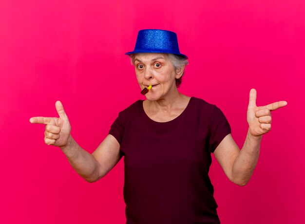 Surprised elderly woman wearing party hat points at sides blowing whistle on pink
