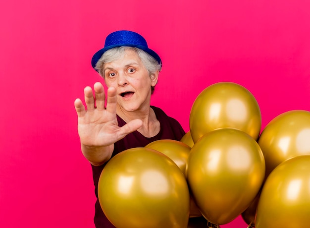 Surprised elderly woman wearing party hat standing with helium balloons stretching out hand on pink