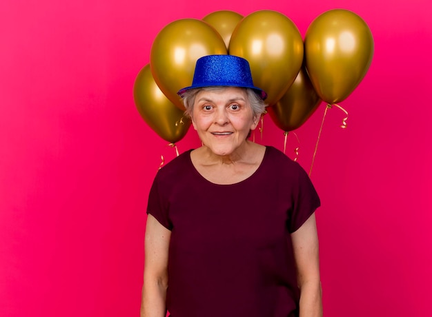 Donna anziana sorpresa che indossa il cappello del partito si trova di fronte a palloncini di elio che guarda l'obbiettivo sul rosa