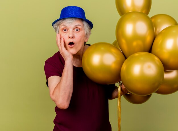 Удивленная пожилая женщина в партийной шляпе стоит с гелиевыми шарами, положив руку на лицо, изолированное на оливково-зеленой стене с копией пространства