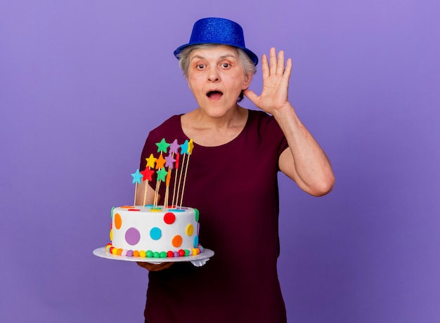 Surprised elderly woman wearing party hat stands with raised hand holding birthday cake isolated on purple wall with copy space