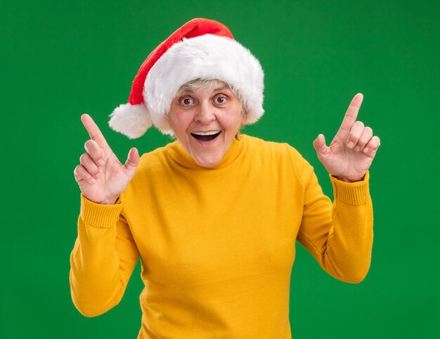 Surprised elderly woman with santa hat pointing up isolated on purple background with copy space