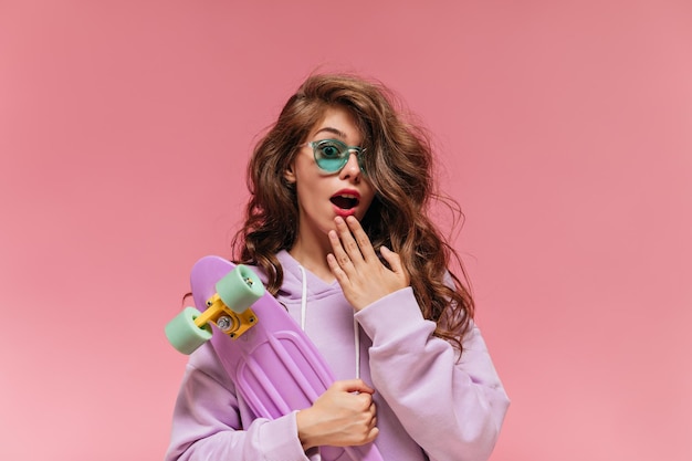 Surprised emotional curly woman holds colorful longboard Pretty girl in purple hoodie and green sunglasses looks into camera on pink background