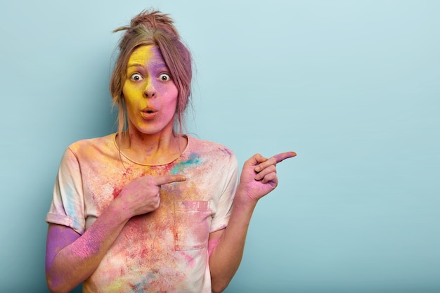 surprised emotional female model has bated breath, dirty with colorful powder, has multicolored face, demonstrates something on blank space. Holi festival celebration concept.