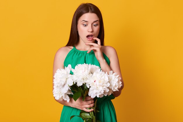 Surprised emotional lady looking at bunch of white peonies, opening her mouth and eyes widely, touching her cheek with finger, being impressed by spring present, guessing whose gift it must be.