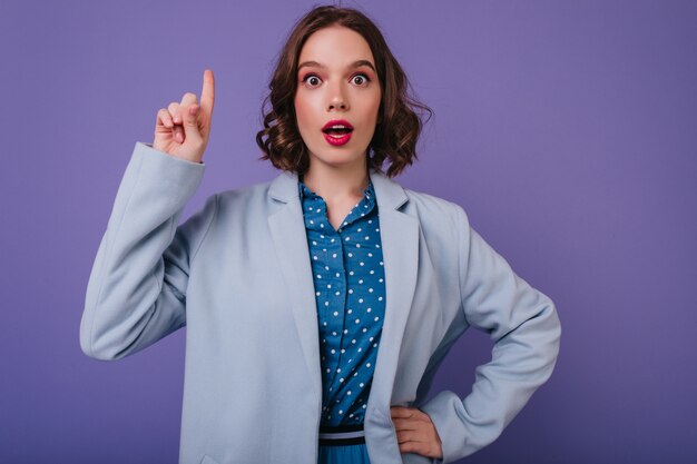 Surprised enchanting woman with curly hairstyle standing on purple wall. Indoor shot of charming european girl expressing amazement.