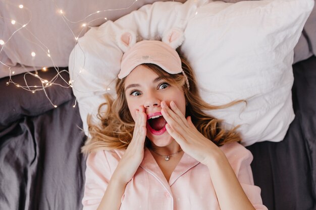 枕の上に横たわっているピンクのアイマスクで驚いた魅惑的な若い女性。朝のベッドでポーズをとっているブロンドの髪を持つ驚いた女の子のオーバーヘッドの肖像画。