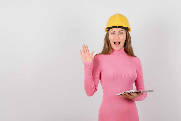 Surprised engineer girl is rasing up her left hand by holding tablet computer on white background