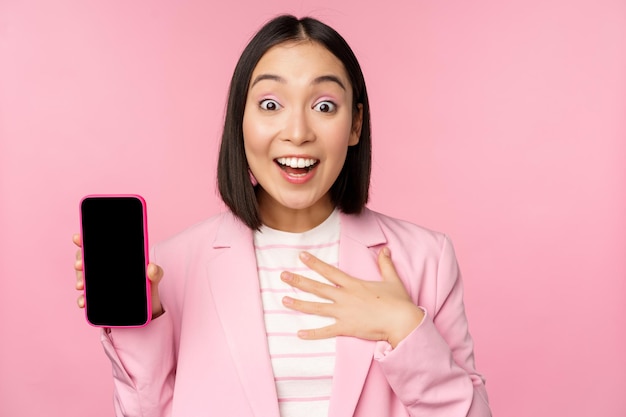 Удивленная восторженная азиатская деловая женщина показывает интерфейс приложения смартфона на экране мобильного телефона, стоящего на розовом фоне