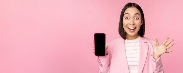 Surprised enthusiastic asian businesswoman showing mobile phone screen smartphone app interface standing against pink background