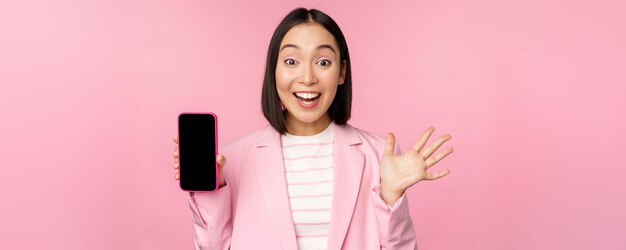 Surprised enthusiastic asian businesswoman showing mobile phone screen smartphone app interface standing against pink background