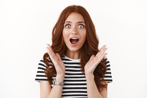 Surprised enthusiastic cute, ginger girl in striped t-shirt hearing excellent news, open mouth excited and impressed, gasping thrilled, stare camera at breathtaking copy space, white wall