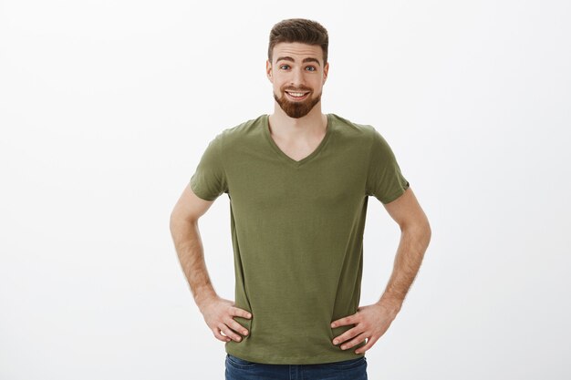 Surprised and enthusiastic good-looking athletic bearded guy in t-shirt holding hands on hips and smiling raising eyebrows from amusement and amazement posing relaxed over white wall