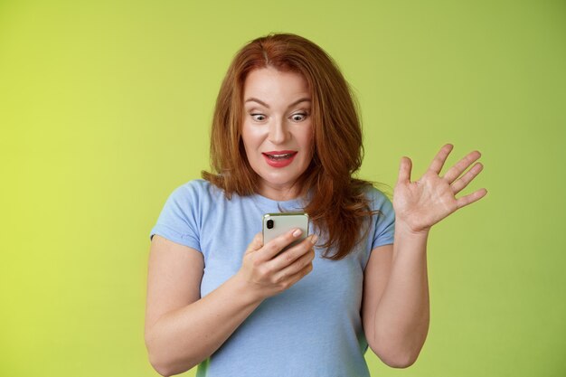 Surprised enthusiastic happy redhead mature middleaged woman receive excellent message read perfect news social media hold smartphone stare astonished pleased mobile phone screen raise hand triumph