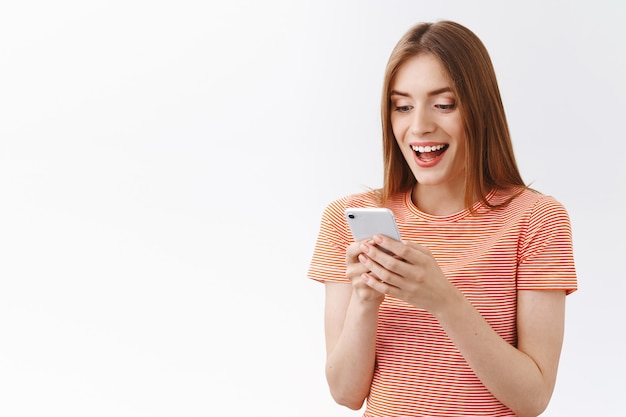 Sorpresa, entusiasta, bella giovane donna in t-shirt a righe, tenere in mano lo smartphone leggere notizie incredibili, aprire la bocca affascinata, sorridere reagisce a prezzi meravigliosi sul sito di shopping di app per dispositivi mobili