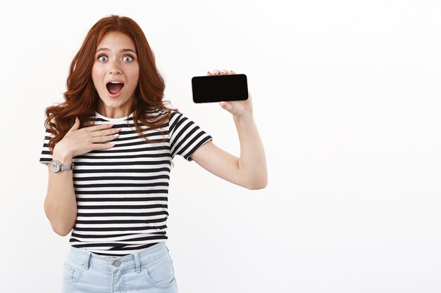 Surprised enthusiastic young female redhead gamer look speechless and impress, cannot realise she bit record, showing smartphone display horizontally, gasping with awe and amazement