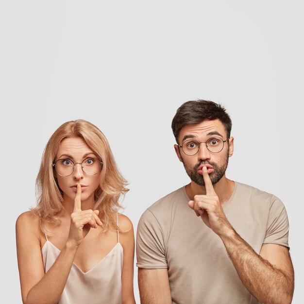 Surprised European woman and man makes silence gesture, tells confidential information, ask to be quiet