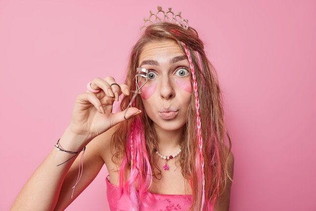 Surprised European woman prepares for party uses eyelashes curler applies makeup beauty hydrogel patches under eyes keeps lips rounded wears crown and festive dress isolated over pink background.