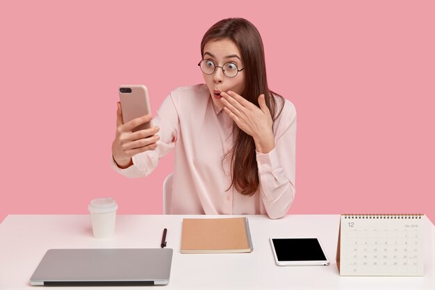 Free photo surprised european young female office worker covers mouth with astonishment, holds cell phone in hand