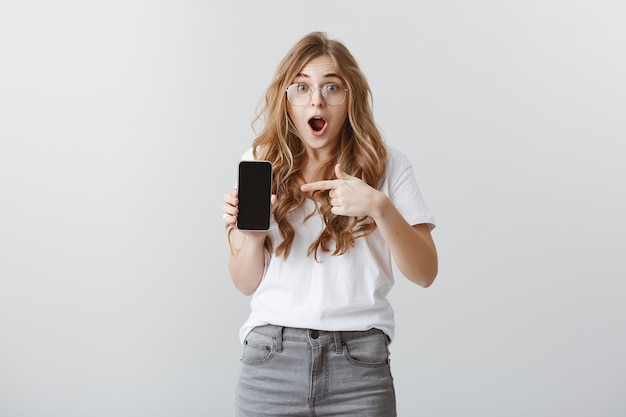 Удивленная и возбужденная блондинка в очках, указывая пальцем на дисплей мобильного телефона, показывая приложение