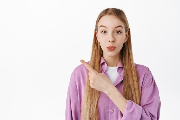 Surprised and excited blond girl pointing aside, showing something interesting with amazed face, telling about annoucement, show advertising, standing over white wall