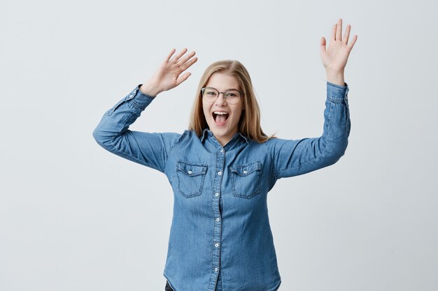 Surprised excited female with blonde hair and spectacles stares , opens mouth widely, raises arms up, being shocked to look at prices at shopping mall, doesn`t believe her eyes.