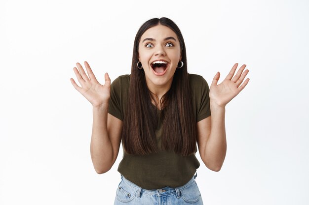 Surprised and excited girl screaming from joy and happy news, raising hands up, staring at front pleasantly astounded, standing against white wall