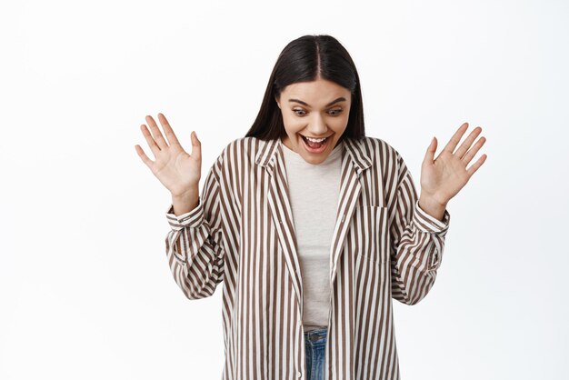 Surprised and excited stylish girl scream from joy and amazement raising hands up amazed looking down and smiling delighted found something on floor white background