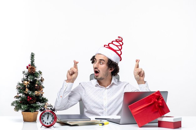 Surprised excited young businessman with funny santa claus hat crossing fingers for luck in the office on white background