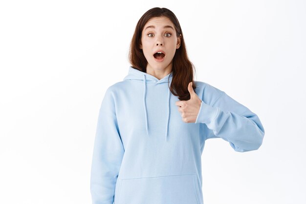 Surprised and excited young woman gasping fascinated showing thumbs up to praise something cool stare at awesome promo offer standing amazed against white background