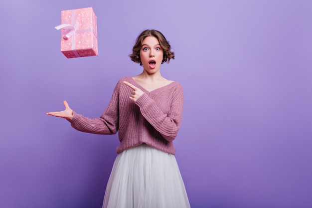 Surprised fascinating lady posing  with purple interior with new year present. Indoor portrait of beautiful girl with short haircut expressing amazement during photoshoot with gift.