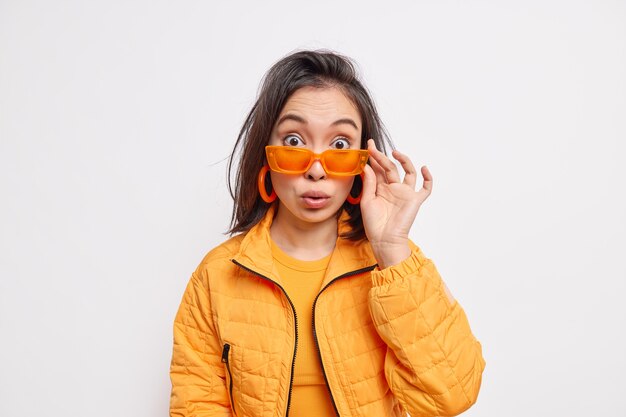 Surprised fashionable brunette young Asian woman wears trendy orange sunglasses jacket and earrings reacts on something astonishing isolated over white wall. Style and fashion concept.