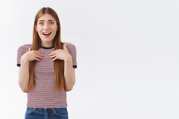 縞模様のTシャツを着た驚きとお世辞の楽しいヨーロッパの若い女の子、手のひらを胸に押して、娯楽と同情からため息をつき、喜んで笑って、素晴らしい贈り物を受け取り、白い背景に立つ