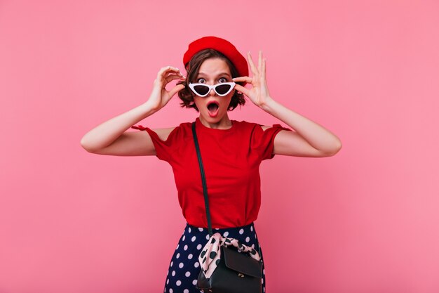 Surprised french girl in stylish sunglasses posing. Indoor photo of elegant white woman in red clothes.