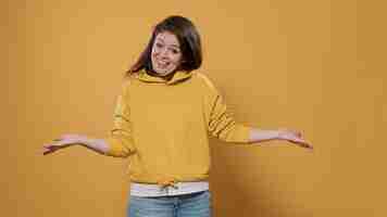 Free photo surprised friendly woman doing greeting gesture smiling and welcoming person in studio. casual young adult wearing hoodie with positive attitude being expressive, joyful, sociable doing hand salute.