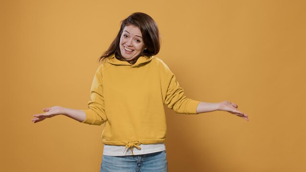 Surprised friendly woman doing greeting gesture smiling and welcoming person in studio. Casual young adult wearing hoodie with positive attitude being expressive, joyful, sociable doing hand salute.