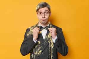 Free photo surprised funny man adjustes bowtie dressed in formal outfit wears round spectacles dresses for special occasion smeared with serpantine spray isolated over yellow background preparing for holiday